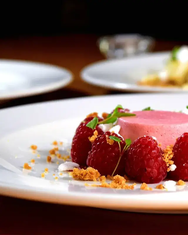 Dessert dressé sur une assiette blanche : mousse rose entourée de framboises et d'éclats croustillants