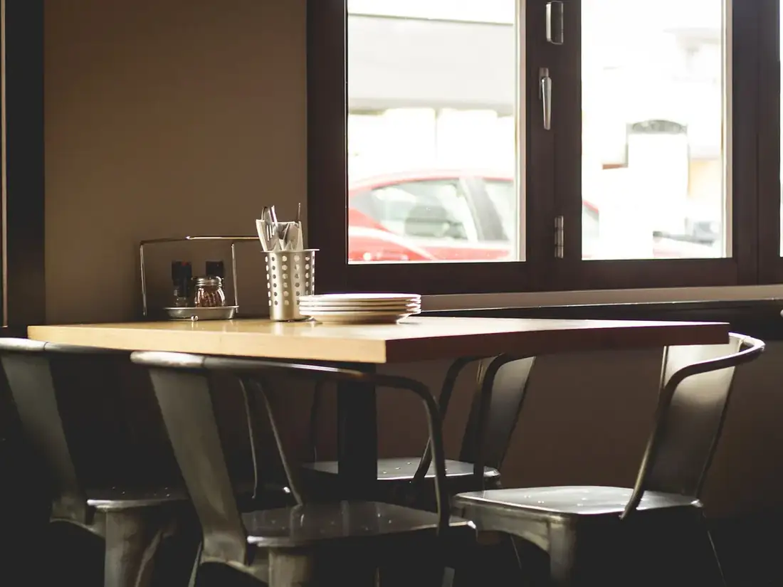 Coin de salle de restaurant avec une table en bois clair et des chaises métalliques près d'une grande fenêtre