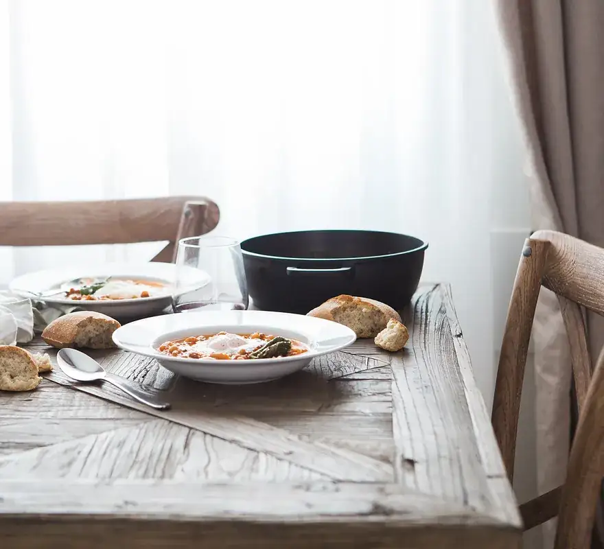 Table en bois clair près d'une fenêtre, avec deux assiettes creuses, du pain et un verre de vin rouge