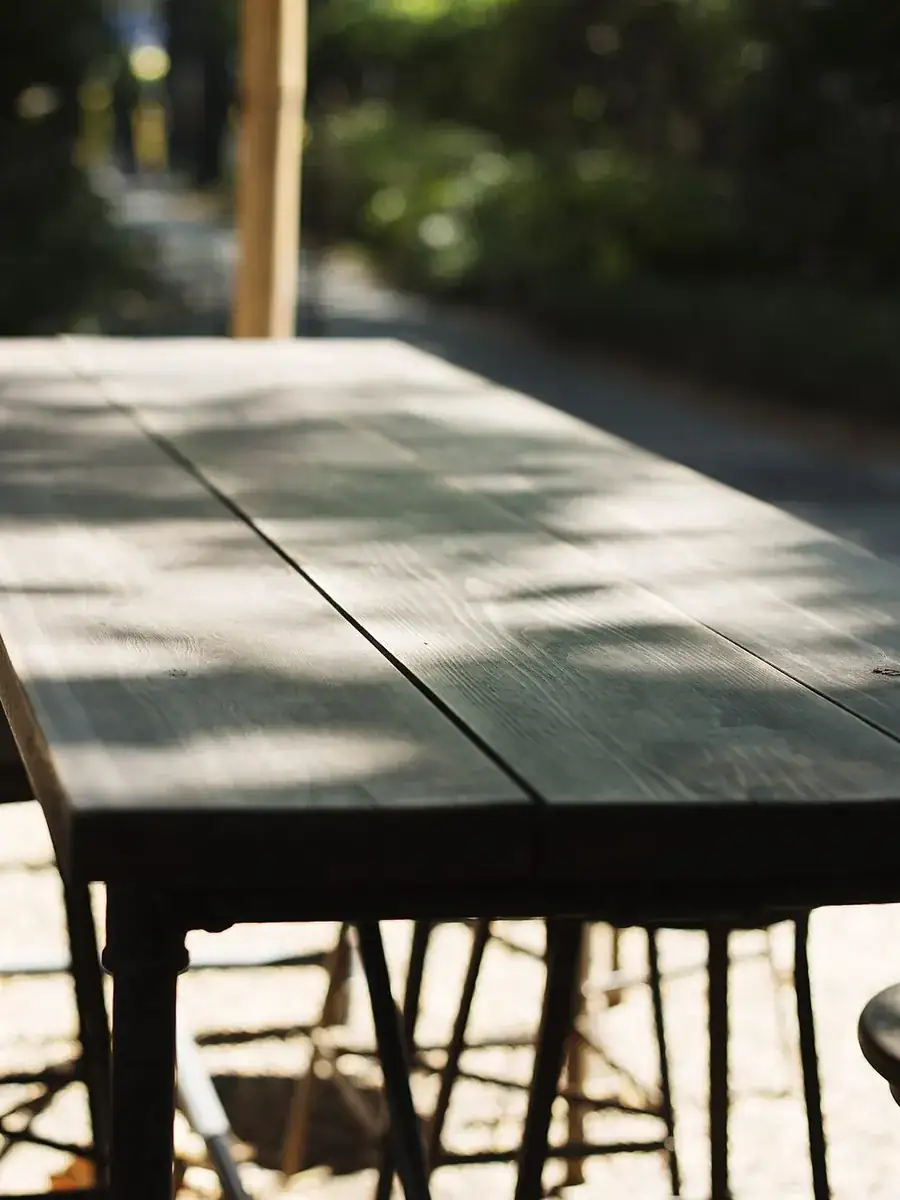 Longue table en bois et tabourets sur une terrasse ombragée, éclairée par le soleil